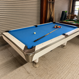 A white and silver pool table with a bright blue felt surface, shown in a showroom or garage setting with a striped billiard rack and another green table in the background.