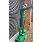A full-body superhero statue or collectible figure wearing a green and black suit, posed with one arm extended, standing on a green display base outdoors beside a metal roll-up door and brick wall.