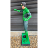 A green and gray superhero statue of a man in a dynamic pose, standing on a green base outdoors in front of a gray metal shutter.