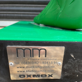 Close-up of a metal brand plate on a green and black machine, with the text 'mm' and 'OXMOX' visible, set against a background of gravel and a green plastic object.